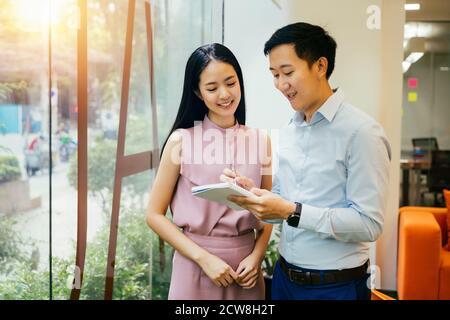 Asian businessman and woman smiling and working together, writing in notebook, collaboration, cooperation, teamwork Stock Photo