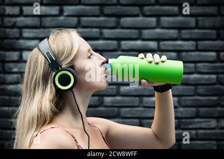 Young blond woman on black background drink fresh spring water from green reusable bottle. Healthy lifestyle in summer city. Stock Photo