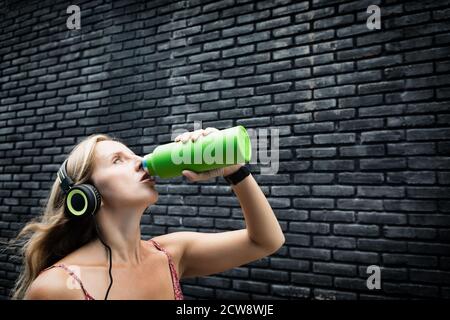 Young blond woman on black background drink fresh spring water from green reusable bottle. Healthy lifestyle in summer city.