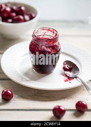 White ceramic bowl of cherries and homemade jam in jar on white rustic wooden table. Close up wih copy space. Stock Photo