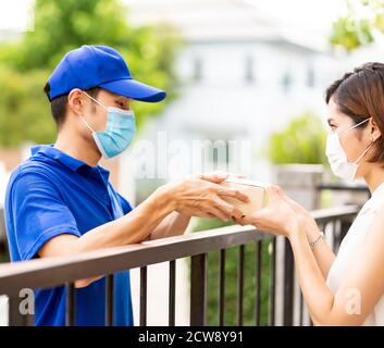 Asian deliver man in blue shirt handling package and give to young woman costumer at house. They wear face mask to reduce coronavirus COVID-19 spreadi Stock Photo