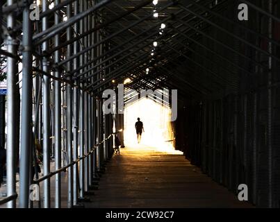 Man walking down the city sidewalk under construction scaffolding with a bright light glowing in the background