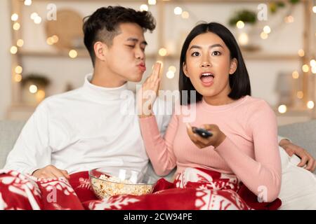 Excited Asian Wife Watching TV Refusing To Kiss Husband Indoors Stock Photo