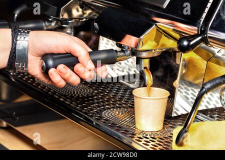 The process of making aromatic coffee, the barista holds the handle of the coffee machine in his hand and pours coffee into a cardboard cup. Stock Photo