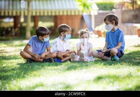 Young children in protective masks on faces outdoors. Quarantine. Kids wearing safety masks while sitting on grass in park. Coronavirus prevention. Stock Photo