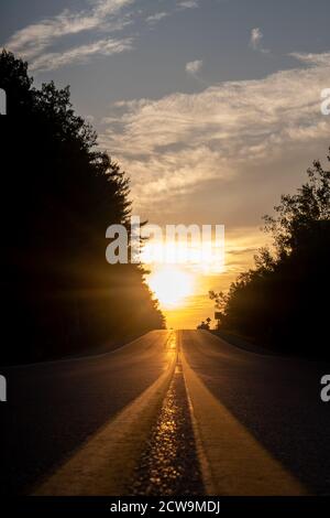 A rural highway is seen from a low angle in a vertical format, with its painted dividing lines leading off into a sunset sky over the horizon. Stock Photo