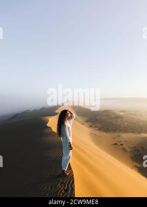 Woman enjoying sunrise on top of huge sand dune. Beautiful warm sun light and mist in morning. Sahara desert, Morocco. Stock Photo