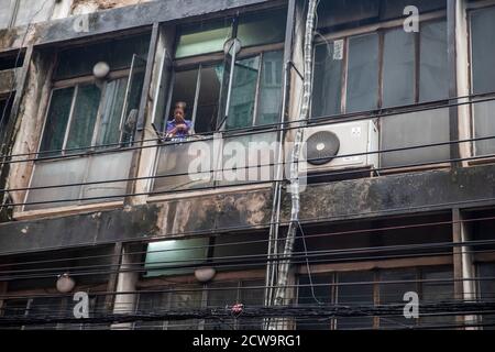 An older woman in a rundown apartment in Bangkok's Chinatown looks out her window at the action on the street below. Stock Photo