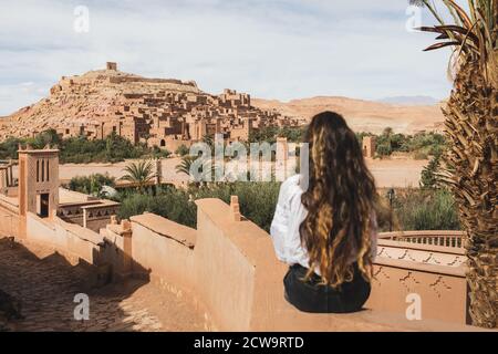 Woman sitting on fence with view of famous moroccan old town ksar Ait-Ben-Haddou. View from behind. Welcome to Morocco, Ouarzazate.