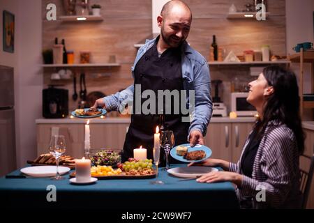 Husband serving wife with tasty food during celebration of relationship in kitchen. Man preparing festive dinner with healthy food, cooking for his woman a romantic dinner, Stock Photo