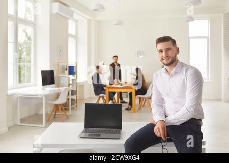 Young businessman with laptop with blank screen sitting on desk in coworking space looking at camera Stock Photo