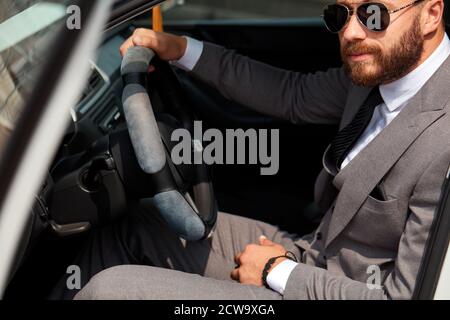 cool businessman in black sunglasses sits inside of car, drive it. successful male in formalwear, in suit. Stock Photo