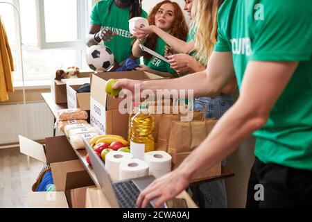 diverse volunteers collecting food and clothes donations in warehouse, multi-ethnic team of volunteers holding donations boxes in a large warehouse, they are putting clothes in boxes Stock Photo