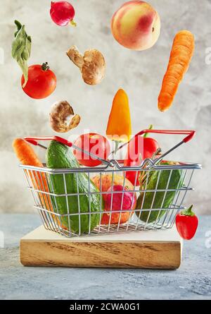 Various vegetables and fruits flying and falling into a supermarket basket standing on a wooden podium. Abundance and delivery concept. Stock Photo