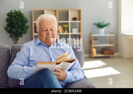 Happy retired man spending leisure at home and reading old book sitting on couch in the living room Stock Photo