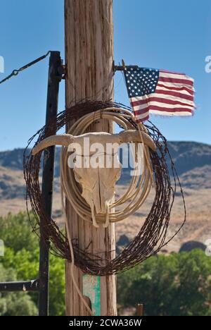 Coil of barbed wire, lasso, bull skull and frayed US flag at ranch gate post in Succor Creek Valley, High Desert Region, Oregon, USA Stock Photo