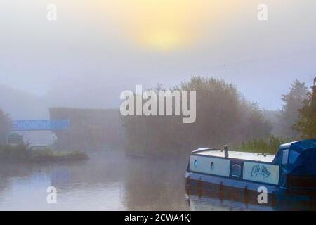 Early morning mist blankets rural Lancashire. Hazy morning as the sun rises over narrowboats at Rufford Marina. When the sun rises, the air and ground warm up. This leads to the air temperature being warmer than the dew point temperature, which causes the fog droplets to evaporate. As the air cools during the longer night the relative humidity increases, which can result in to fog formation. Credit: MediaWorldImages/AlamyLiveNews Stock Photo