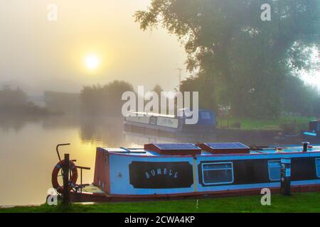 Early morning mist blankets rural Lancashire. Hazy morning as the sun rises over narrowboats at Rufford Marina. When the sun rises, the air and ground warm up. This leads to the air temperature being warmer than the dew point temperature, which causes the fog droplets to evaporate. As the air cools during the longer night the relative humidity increases, which can result in to fog formation. Credit: MediaWorldImages/AlamyLiveNews Stock Photo