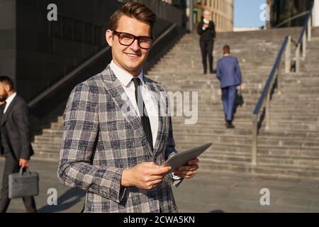 Portrait of young businessman in eyelasses and in elegant suit smiling at camera while using tablet pc outdoors Stock Photo