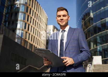 Portrait of young businessman in suit holding folders with documents and smiling at camera standing in the city Stock Photo