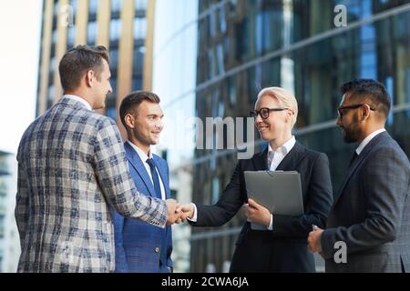 Businessman and businesswoman shaking hands and greeting each other while standing in the city outdoors with their colleagues Stock Photo