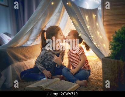 Two cute little children are reading a book with flashlights in tent. Happy girls playing at home. Funny lovely kids having fun in room. Stock Photo