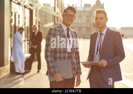 Portrait of two young businessmen with documents and digital tablet smiling at camera while standing on city street Stock Photo