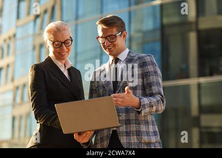 Two business colleagues watching on monitor of laptop and using it in their work while standing outdoors Stock Photo