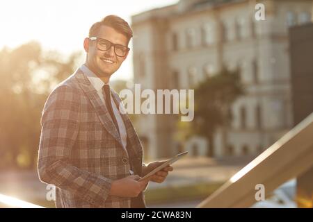 Portrait of young businessman in eyeglasses holding digital tablet and smiling at camera outdoors Stock Photo