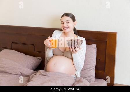 Pregnant woman enjoys eating crunchy chocolate cereal balls from a bowl and drinking juice relaxing in bed. Dieting during pregnancy concept. Stock Photo