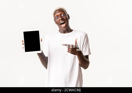 Portrait of excited and amazed african-american blond guy, looking impressed and pointing finger at digital tablet screen, standing white background Stock Photo
