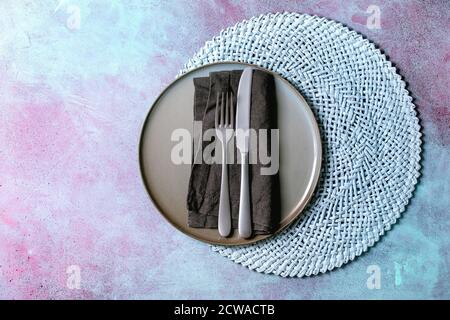 Empty ceramic plate with fork and knife on textile napkin on knitted round napkin over blue concrete background. Flat lay, space Stock Photo