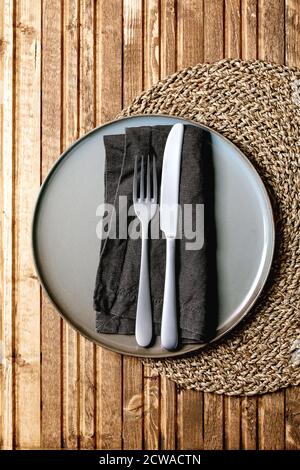 Empty ceramic plate with fork and knife on textile napkin on knitted straw round napkin over wooden plank background. Flat lay, space Stock Photo