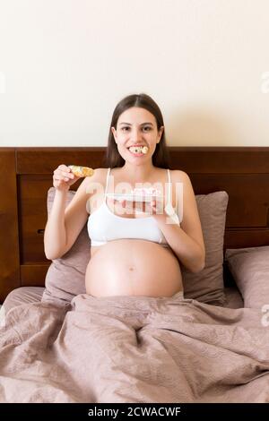 Pregnant woman is eating many donuts relaxing on the sofa. Unhealthy dieting during pregnancy concept. Stock Photo