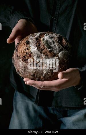 Man in black shirt holding in hands fresh baked artisan round homemade chocolate and cranberries rye bread. Stock Photo