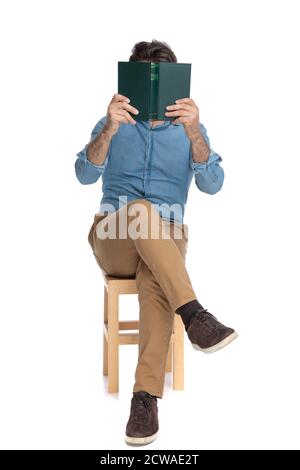 Old casual man covering his face with a book while sitting on a chair on white studio background Stock Photo