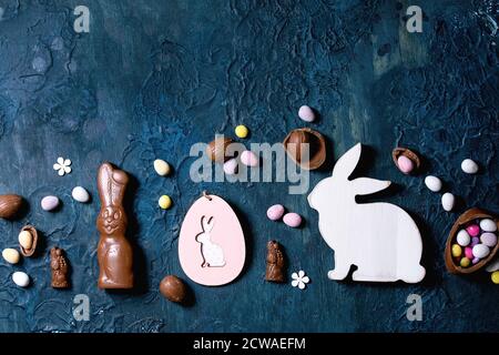 Easter greeting card with wooden rabbit decorations, chocolate sweets, bunny and eggs on classic blue texture background. Flat lay, copy space. Stock Photo