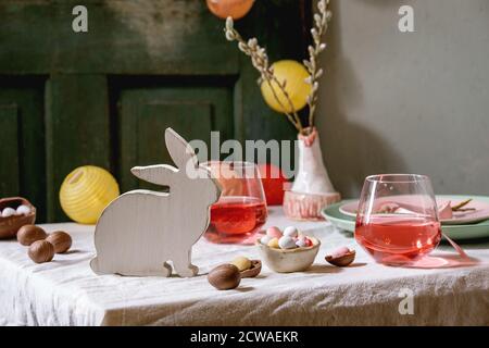 Easter table setting with wooden rabbit decoration, chocolate eggs and sweets, glasses of pink cocktail drinks, empty plates on white tablecloth. Ligh Stock Photo