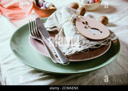 Easter table setting with wooden rabbit decoration, chocolate eggs and sweets, glasses of pink cocktail drinks, empty plates with textile napkin, cutl Stock Photo
