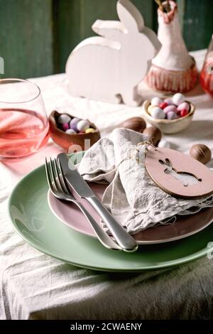 Easter table setting with wooden rabbit decoration, chocolate eggs and sweets, glasses of pink cocktail drinks, empty plates with textile napkin, cutl Stock Photo