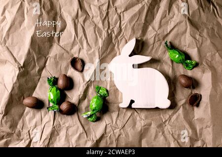 Easter greeting card with wooden rabbit decorations, chocolate sweets and eggs on crumpled craft paper background. Happy Easter inscription. Flat lay, Stock Photo