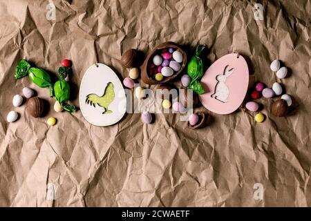 Easter greeting card with wooden rabbit and chicken decorations, chocolate sweets and eggs on crumpled craft paper background. Flat lay, copy space. Stock Photo