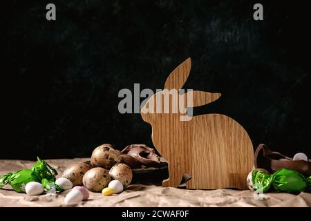Easter mood still life with wooden rabbit decoration, golden quail eggs, traditional chocolate eggs and sweets on table with crumpled craft paper. Dar Stock Photo