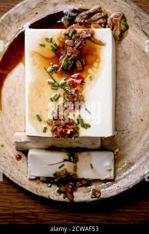 Silk tofu japanese soy cheese whole piece with chili ginger, chive and soy sauce topping on ceramic plate over wooden table. Flat lay, close up Stock Photo