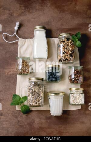 Homemade crunchy puffed millet grain granola with dried fruits and nuts, yogurt, milk, muesli in different glass jars on cotton bag. Brown background. Stock Photo