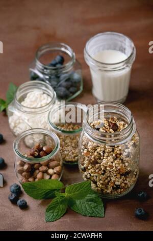 Homemade crunchy puffed millet grain granola with dried fruits and nuts, yogurt, milk, muesli in different glass jars, mint. Brown background. Healthy Stock Photo