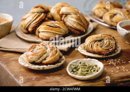 Traditional Swedish cardamom sweet buns Kanelbulle on plate, ingredients in ceramic bowl above on wooden table. Stock Photo