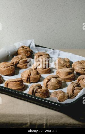 Ready to bake traditional Swedish cinnamon sweet buns Kanelbulle on oven tray cover by baking paper on grey linen table cloth. Stock Photo