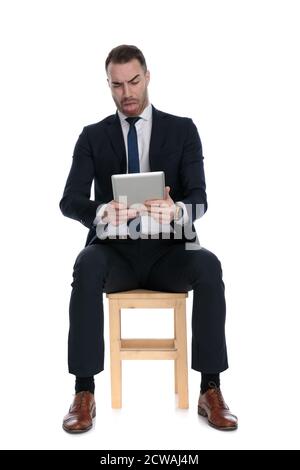 Disgusted businessman holding tablet, frowning with his tongue exposed while sitting on a chair on white studio background Stock Photo