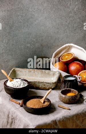 Ingredients for home baking. Flour, eggs, cane sugar, milk in different ceramic bowls, blood oranges and spices on linen table cloth. Grey texture bac Stock Photo
